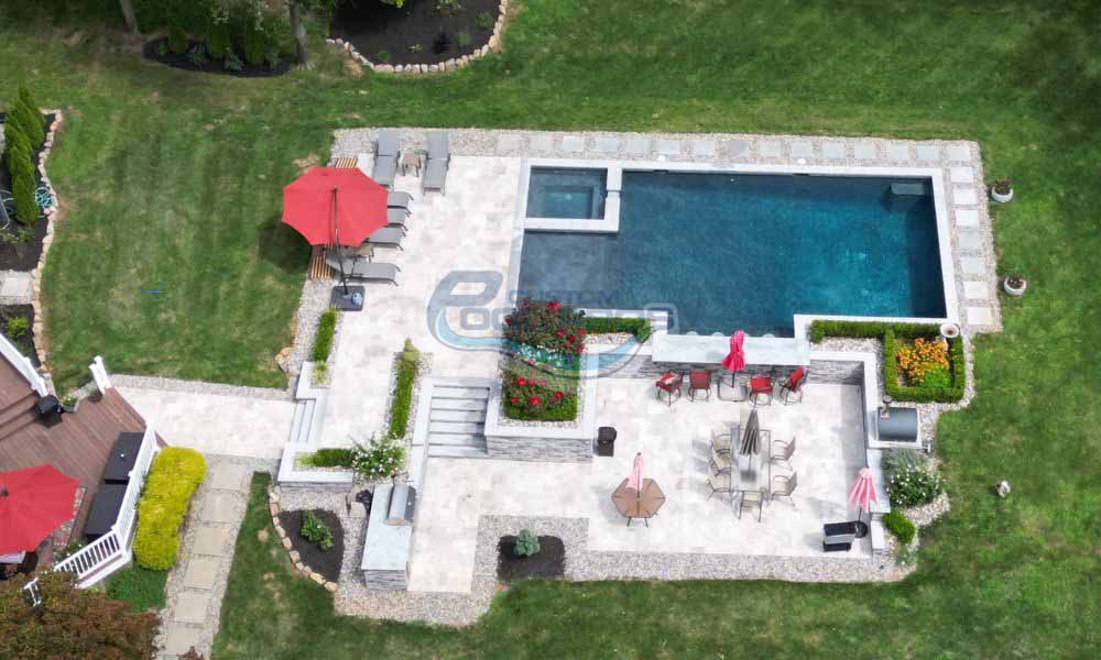 Aerial view of a backyard with a rectangular swimming pool, lounge chairs, red umbrellas, tables, patio areas, green lawn, and surrounding landscaping, landscape installation