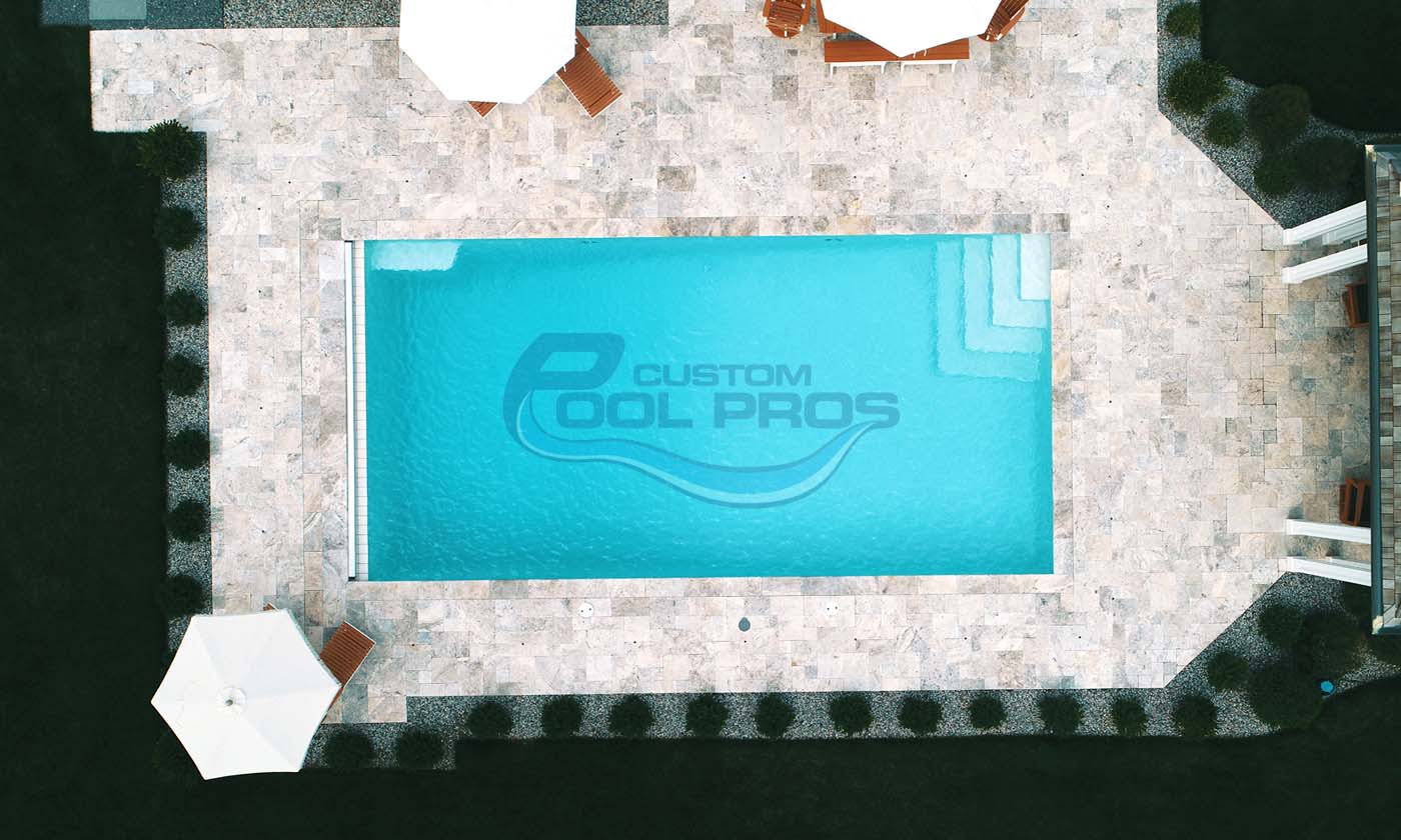 Aerial view of a custom swimming pool with clear blue water, surrounded by stone decking and lounge chairs, featuring the logo of Custom Pool Pros.