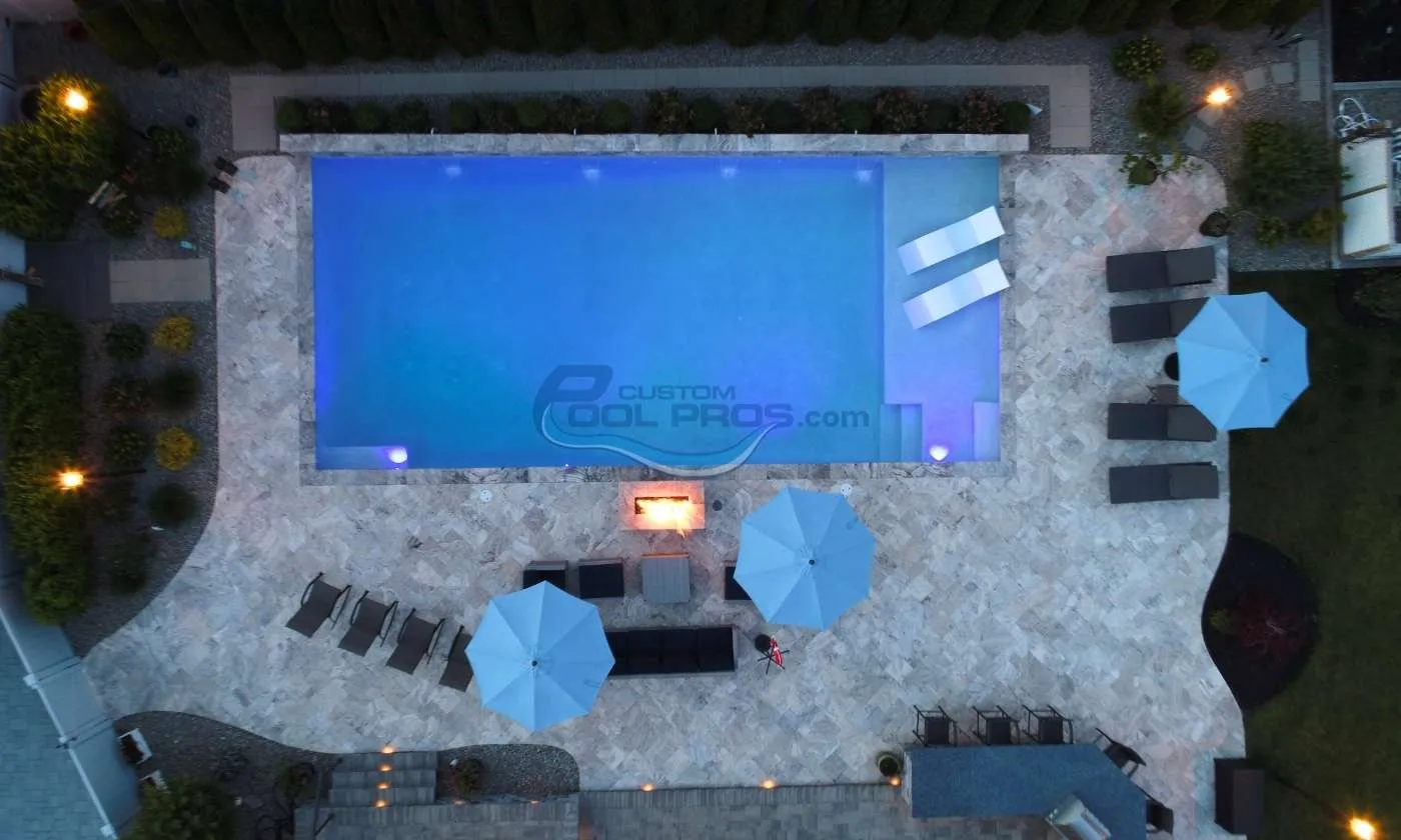 Aerial view of a modern swimming pool surrounded by stone tiles, lounge chairs, and blue umbrellas, featuring glowing lights and a cozy fire pit area, best pool companies in nj